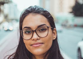 A confident woman wearing glasses smiles subtly while standing on an urban street with blurred background.